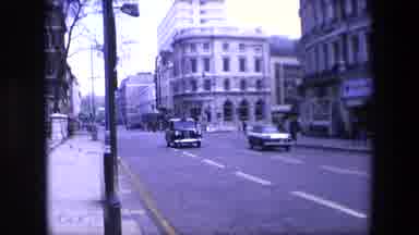 London England (1967): Two Cars Driving Through City Street And People Walking In