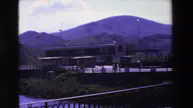 Loudes France (1969): People Walking Away From A Far Off Building By The Mountains