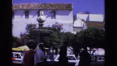Spain (1972): People Walking Towards Buildings In A City With Cars Parked Nearby