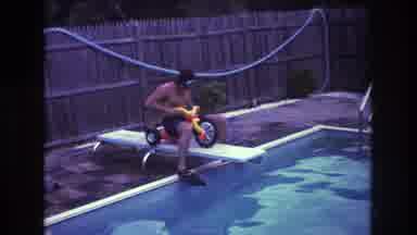 Boston USA (1975): A Man Rides A Tricycle Off Of A Diving Board Into A Swimming