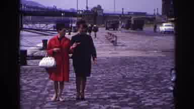France (1967): Two Women Link Arms And Cross A Cobblestone Street Together
