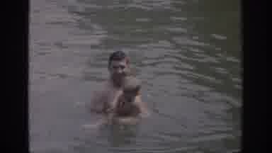 Oklahoma USA (1967): Man Helps Small Boy Stay Afloat On Water Raft In Lake