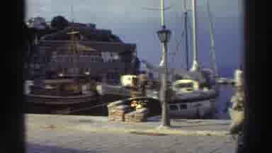 Greece (1976): Three People Walking Toward Sailboats Docked At A Seaside Marina