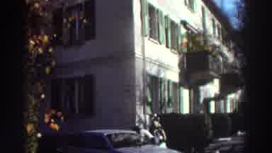 GREECE-1976: One Person Filming a Sunlit Street with a Classic Car and Mountain View