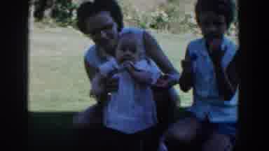 Michigan USA (1958): A Woman Holds A Baby On A Lawn While Three Little Girls Sit