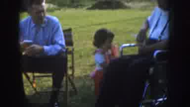 Nebraska USA (1955): Toddler Walking About Some Loungers Sitting In Chairs In An