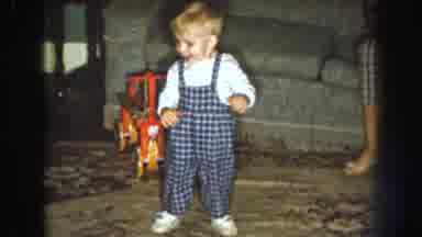 Nebraska USA (1959): Baby Wearing Blue Overalls Dances On White Rug In Front Of