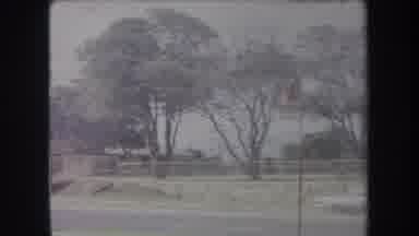 Hawaii USA (1969): White Car Speeding Past A Park With Buildings Nearby