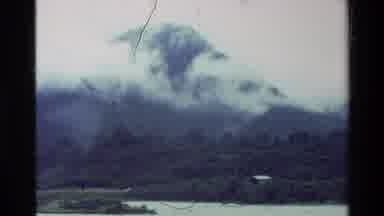 New Zealand (1981): Very High Mountain With A Fog Around It With A Forest Below