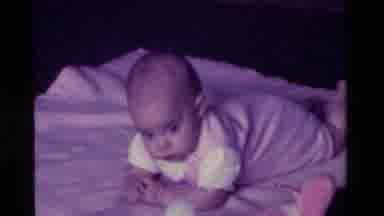California USA (1975): Baby Wearing Pink Overalls Lies On White Blanket Looking