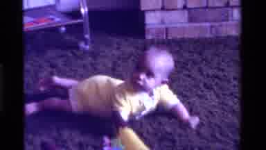 California USA (1975): Baby In Yellow Onesie Lying Prone On Brown Carpet Near