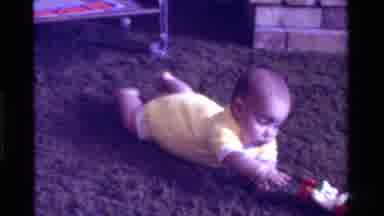California USA (1975): Baby In Yellow Outfit Lies On Belly Kicking And Reaching