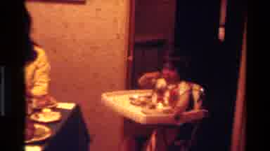 California USA (1977): People And Baby Sitting In Highchair Eating An Evening Meal