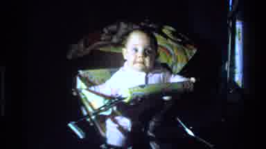 California USA (1977): Baby In A Colorful Stroller Looks Around A Dark Room