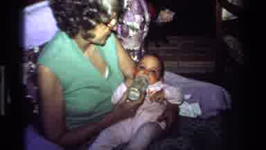 California USA (1977): Grandmother Feeding Baby Girl In Pink Onesie With A Bottle