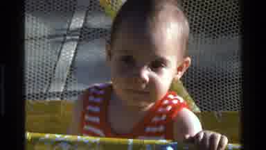 California USA (1977): A Toddler Wearing A Shirt Stands In A Playpen And Chews On