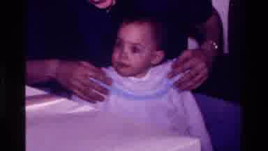 California USA (1977): Baby Sits Alert In An Adult's Lap While Clothing Is