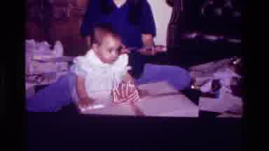 California USA (1977): Baby In A White Dress Crawling Over A Present Wrapped In A