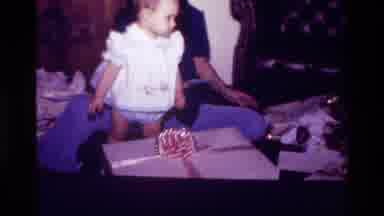 California USA (1977): Adult Playing With A Baby Girl By A Package In A Living