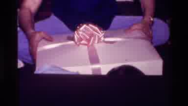 California USA (1977): Person Trying To Open A Present While Baby Plays With It