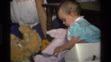 California USA (1977): Baby In A Blue Dress Rummaging Through Toys And Blankets