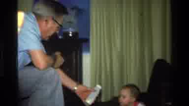 California USA (1977): Older Man Handing A Bottle To A Baby Girl In A Home