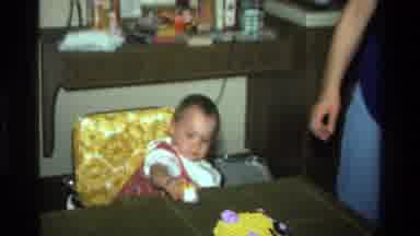 California USA (1977): Baby In Highchair Trying To Eat Part Of A Decorated Cake