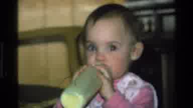 California USA (1977): Baby Wearing White And Pink Dress Drinks From Green Bottle