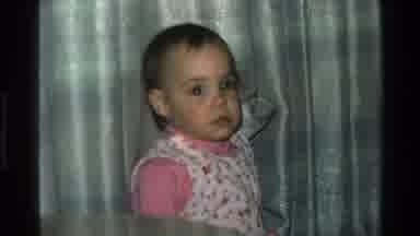 California USA (1977): Baby Girl Looking At The Camera And Then Picking Up A Red