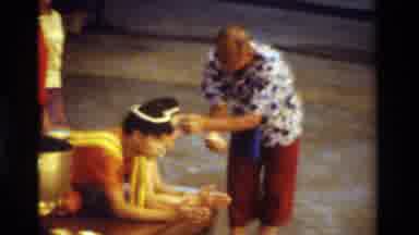 Thailand (1984): A Person Sitting Down While Another Person Puts Something In Her