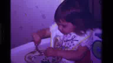 California USA (1979): Baby Girl Sitting In Stool Eating Peas From A Plate With A