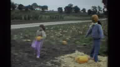 California USA (1984): Girl Carrying Pumpkin Past Scarecrow With Pumpkin Head On