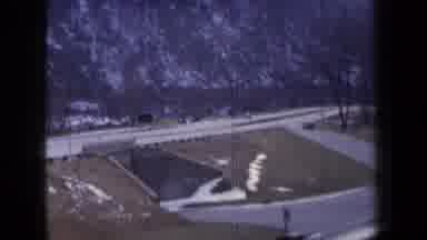 New York USA (1959): Cars Drive Over A Bridge Situated Next To A Snowy Mountain