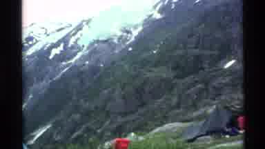 Alaska USA (1977): Mountain Scene With Tents Set Up In A Small Opening Around Tall