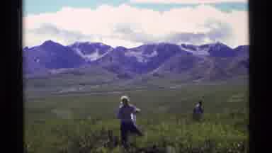 Alaska USA (1977): People Tending To Plants In A Grassy Area With Mountains In The