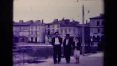 France (1945): Three People Walk Across Bridge Above Water With Houses Behind