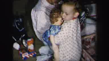 Lynbrook New York (1975): Child Holds Blue Doll While Woman Folds Brown Clothes On