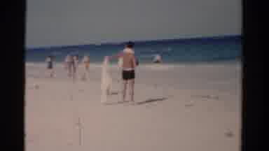 Florida USA (1975): Man Shakes Out Sand From Towel At Beach While A Dog Runs Away