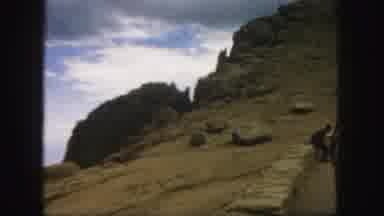 Colorado (1958): Cars And A Dirt Hillside With Large Rocks And Clouds In The
