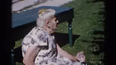 Illinois USA (1962): Older Woman In A White Dress With Blue Flowers Taking A Seat