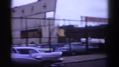 Illinois USA (1962): Old Car Drives Down Street Man Crosses Street In Front Of