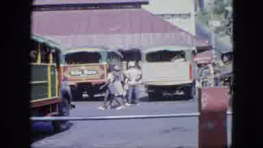 Grenada (1964): People Walking Past Large Green Tour Trucks In Urban Area