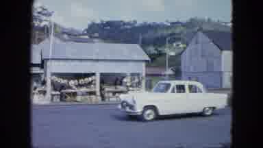 Grenada-1964: Vintage Car Driving Past Market and Warehouse