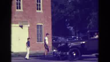 Diller Nebraska (1965): A Man Driving A Nice Car Passing People By A Building