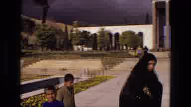 Iran (1972): People Wearing Black Robes Walk Across Courtyard On A Dark Cloudy Day