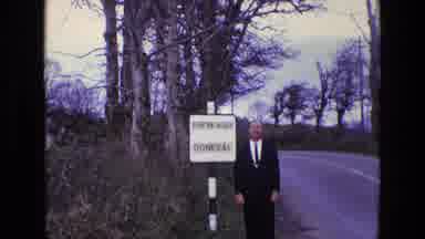 Ireland (1969): Man Standing On Side Of Road Next To Sign Near Trees Smiling