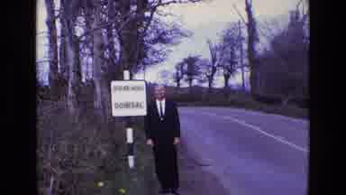 Ireland (1969): Man In Suit Stands By The Road Sign And Waves To The Camera