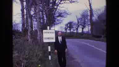 Ireland (1969): Country Road In Farm Land With Man In Suit Waving Near A Sign