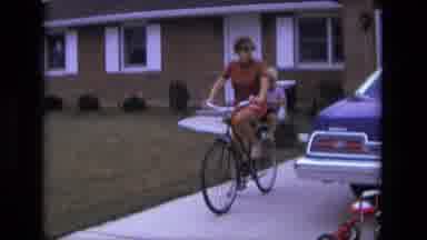 California USA (1975): Woman Takes Off From Driveway On Bicycle With A Child In A