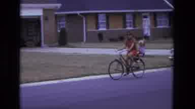 California USA (1975): Mother Rides Bike With Baby In A Seat Attached To The Back
